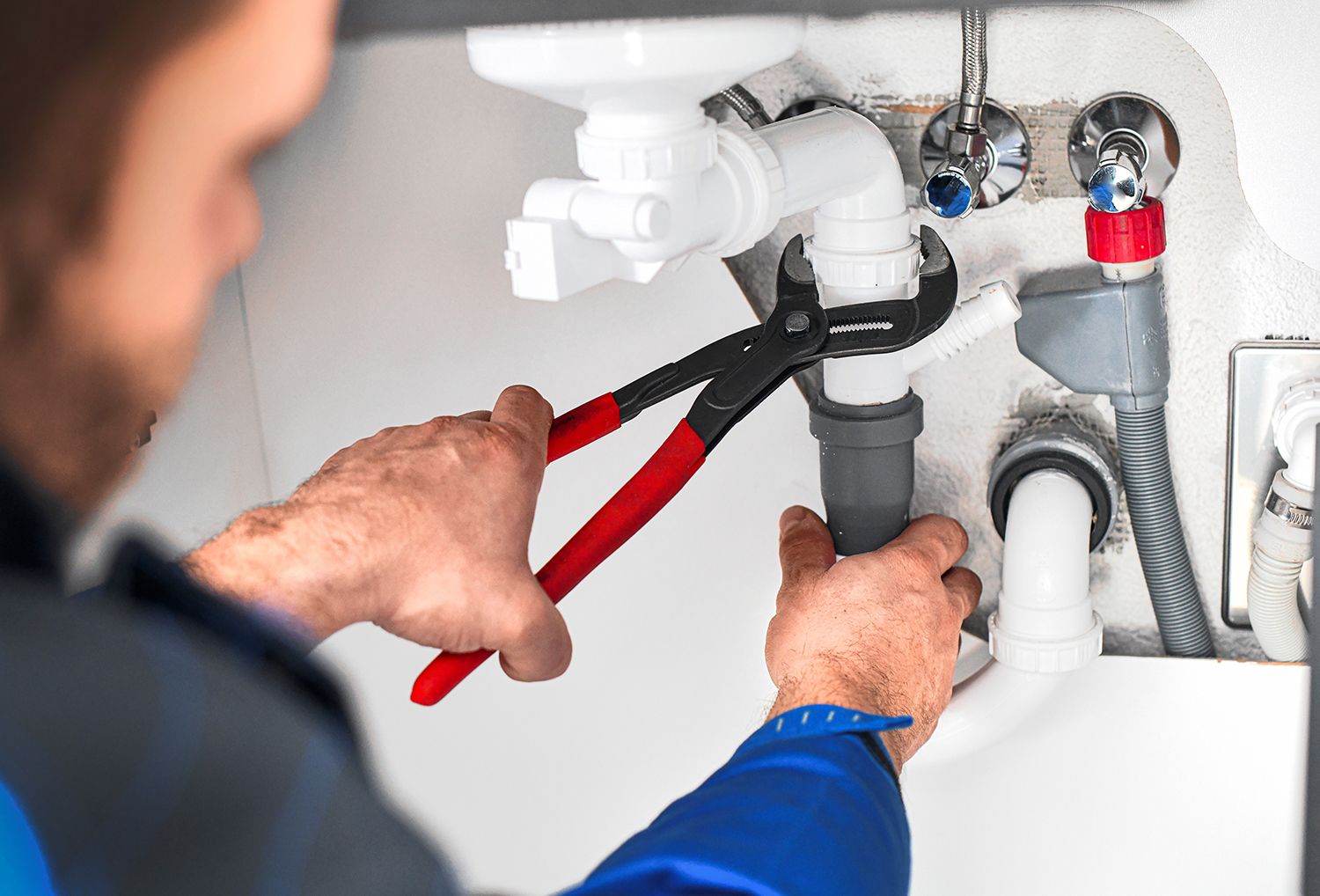 Worker tightening plumbing pipes under a sink using a pair of red-handled pliers.