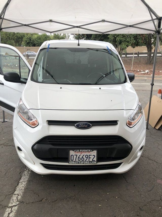 A white ford transit connect is parked under a tent in a parking lot.