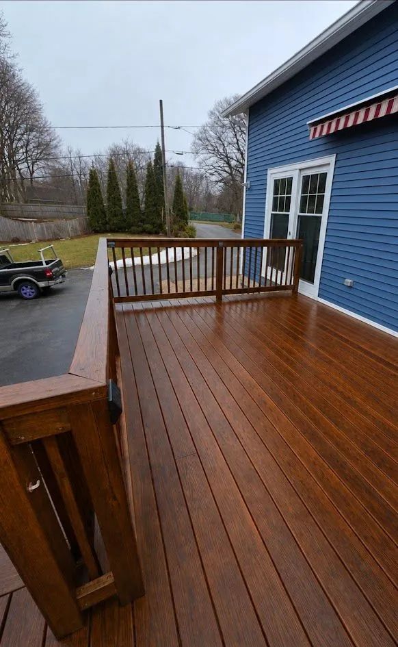 Wet wooden deck beside a blue house, with railing and driveway visible on a rainy day