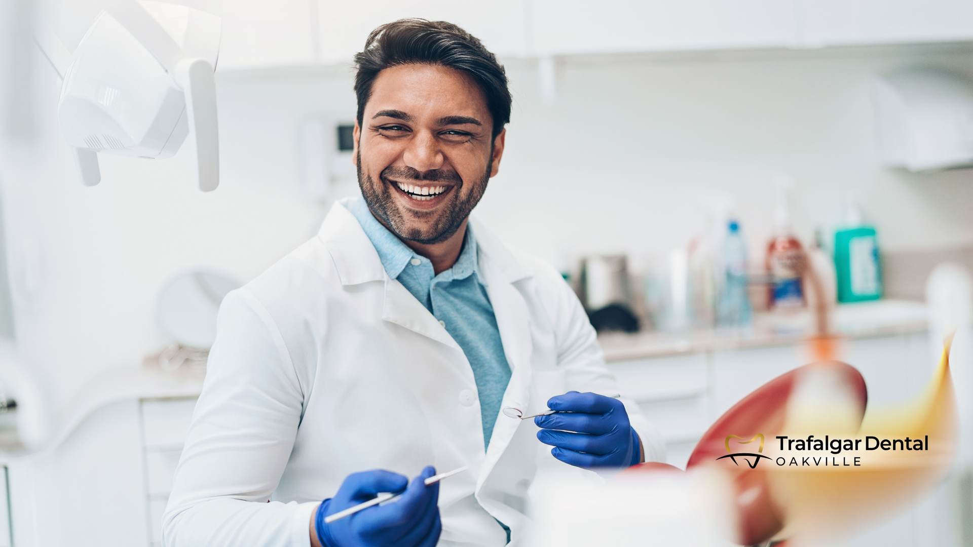 Smiling dentist in white coat and blue gloves, holding tools in a dental office.