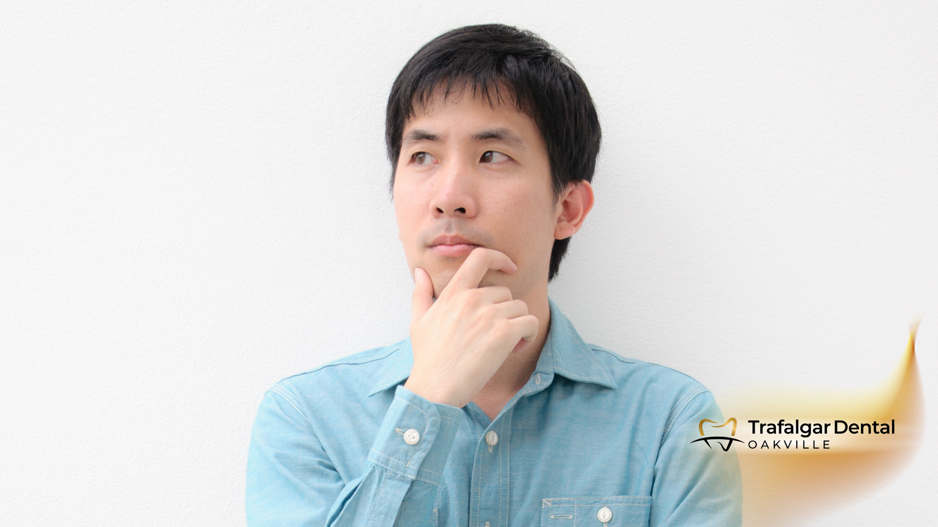 Man in blue shirt, thoughtful expression, hand on chin, standing in front of a white wall.