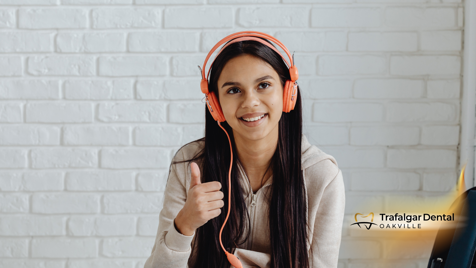Woman with orange headphones gives a thumbs up, smiling. White brick background.
