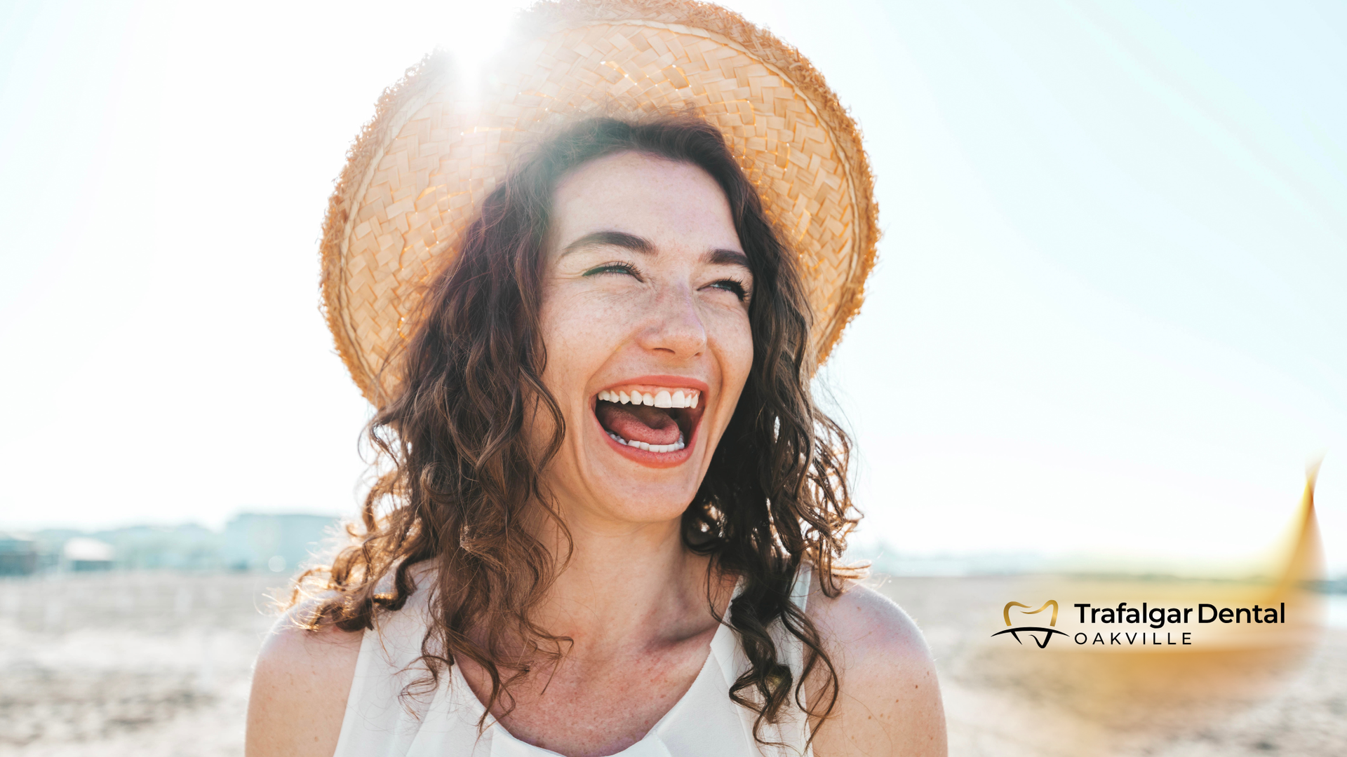 Woman with curly hair and straw hat laughing on a sunny beach.