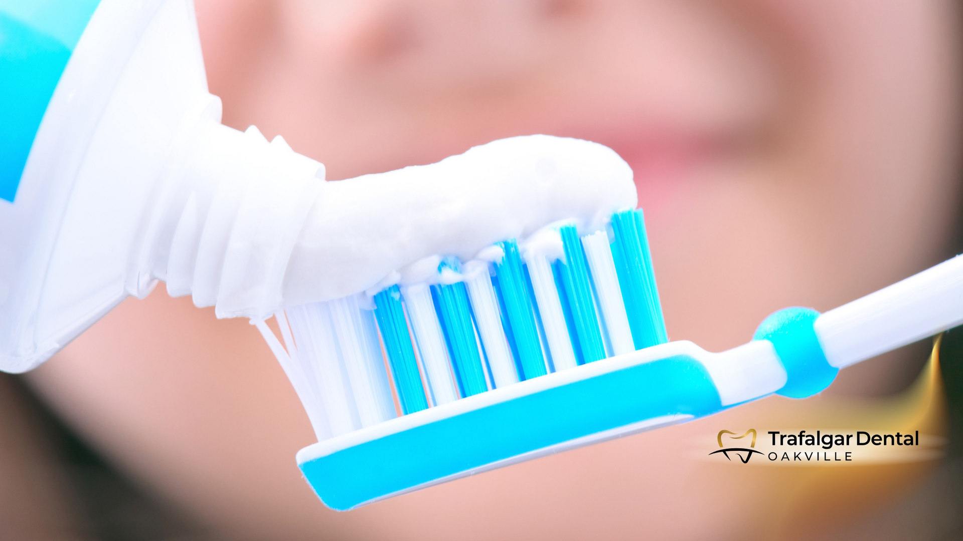 Toothpaste being squeezed onto a blue-bristled toothbrush, with a person's mouth in the blurred background.
