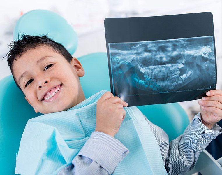 A young boy is sitting in a dental chair holding an x-ray of his teeth.