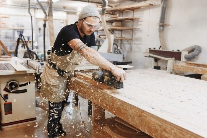 Carpenter planes wood on a workbench in a workshop. He wears safety glasses, a beanie, and apron.