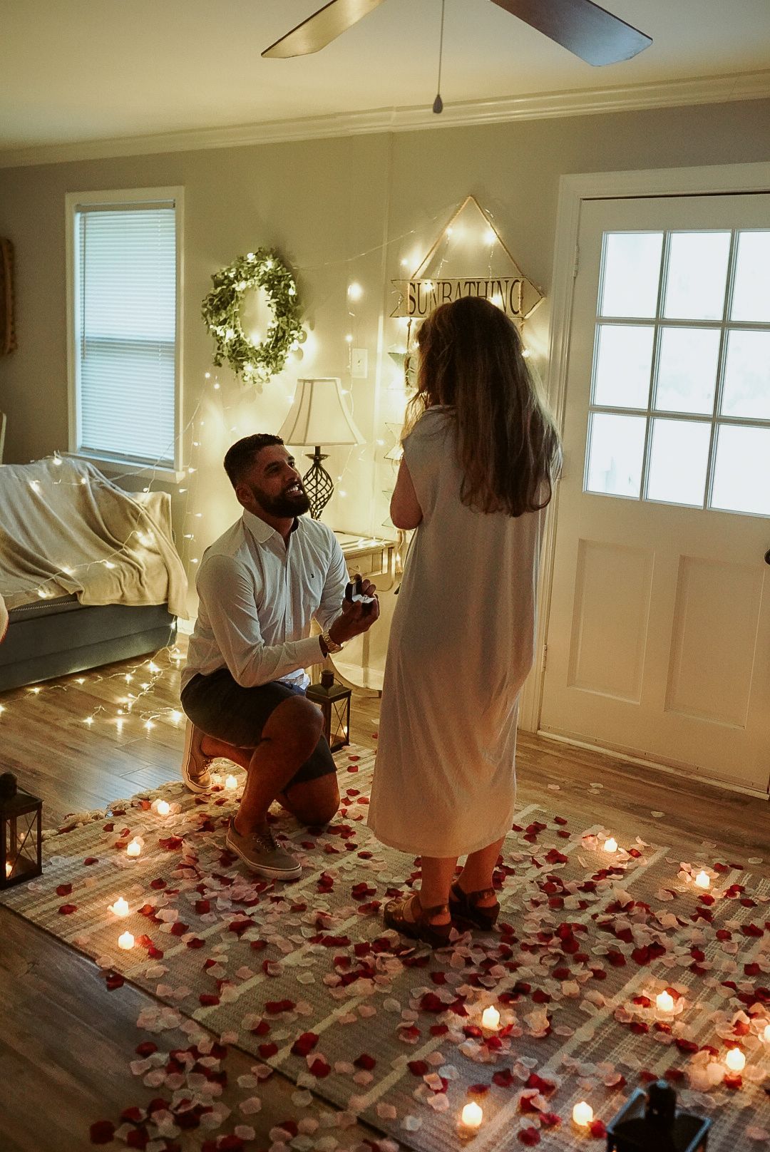A man is proposing to a woman in a living room surrounded by candles and rose petals.