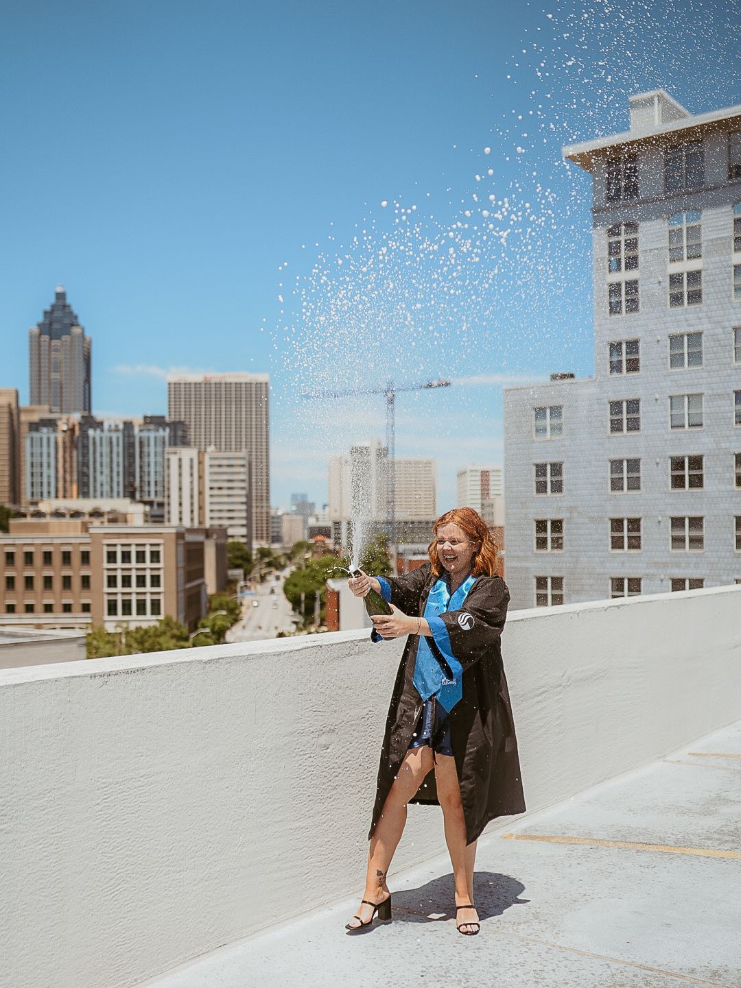 A woman in a graduation cap and gown is standing on a rooftop spraying champagne.