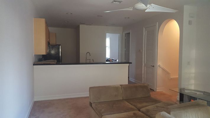 Living room with kitchen in the background; beige couch, white walls, arched doorway leading to stairs.