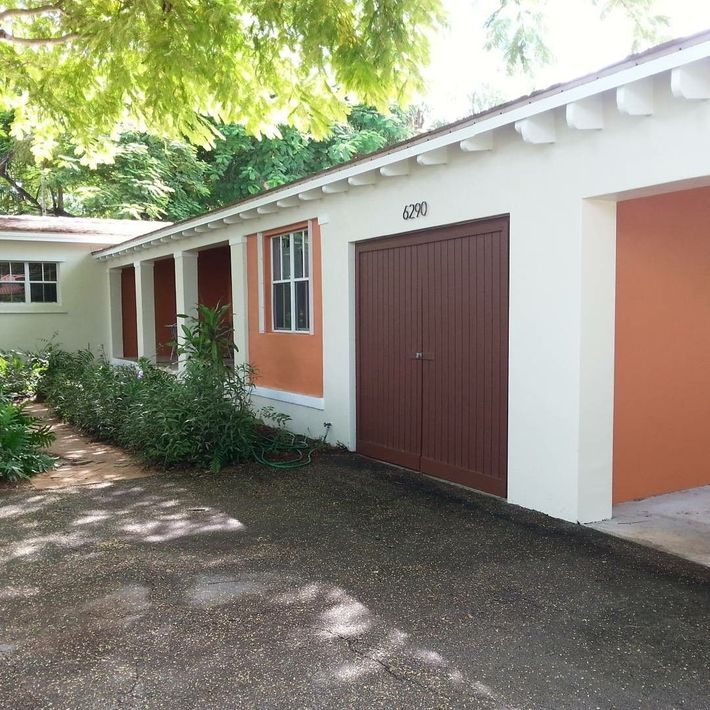 Exterior of a bungalow home, off-white with orange accents, brown garage door, and gravel driveway.