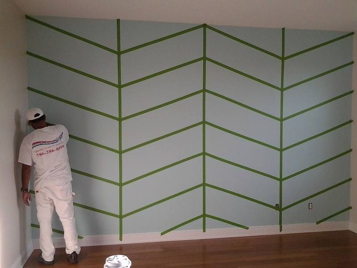 A person tapes off a chevron design on a light blue wall, preparing to paint.