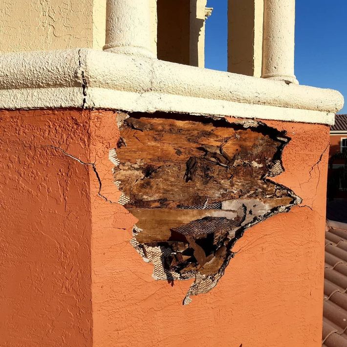Damaged stucco chimney with exposed, rotting wood. Reddish-orange stucco below a cream-colored cap with columns.