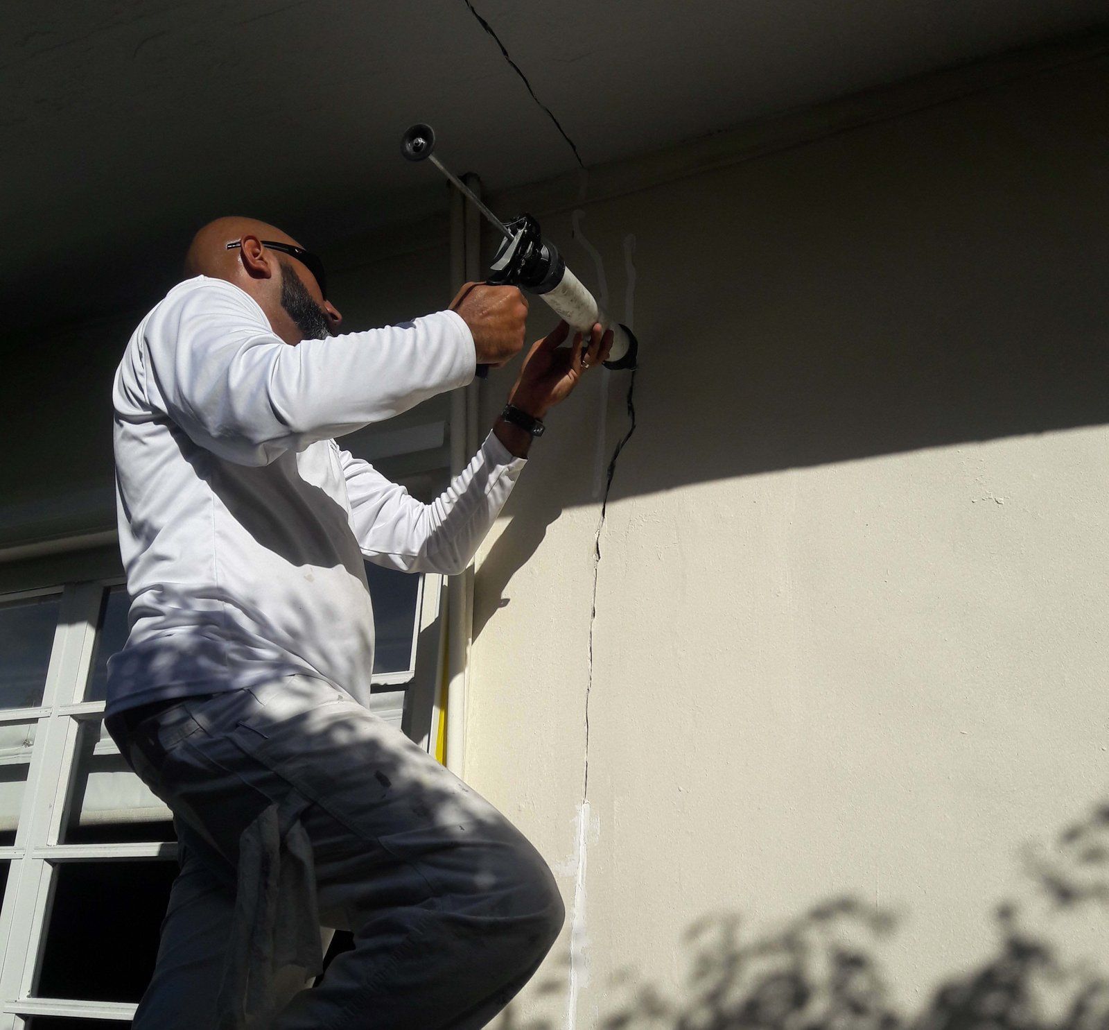 Man on ladder caulking a wall under an overhang. He is wearing a white shirt and pants.