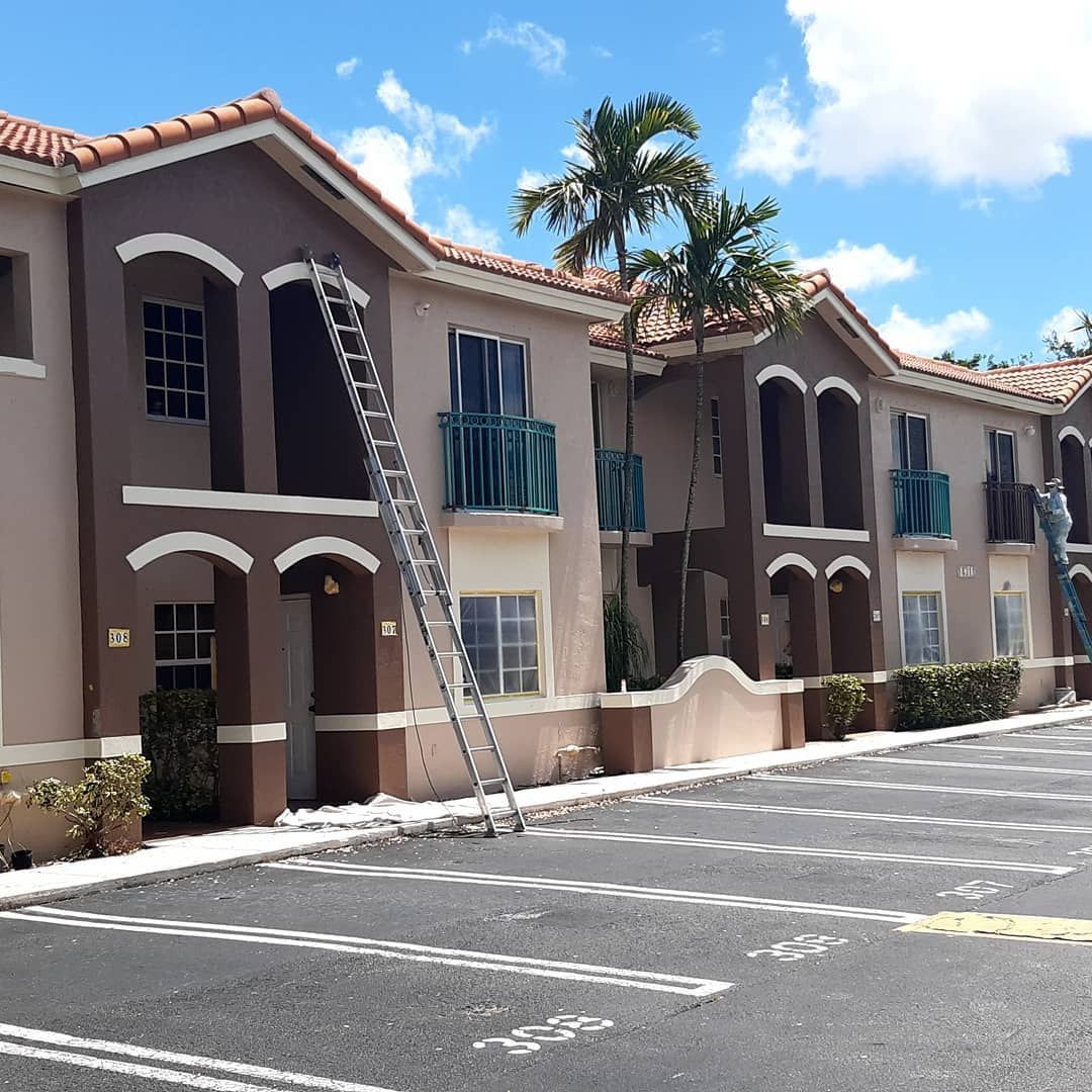 Apartment building exterior with a ladder. Brown and beige walls, teal balconies, blue sky, parking lot.