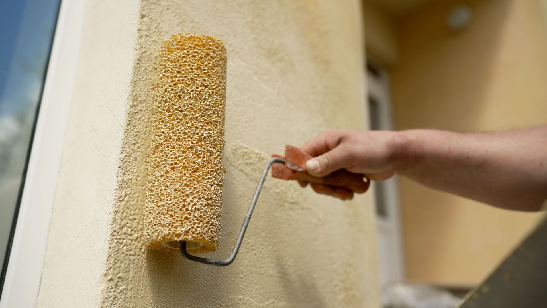 Person using a textured paint roller on a light beige exterior wall.