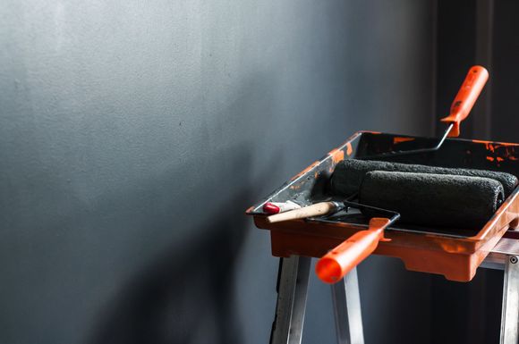 Orange paint tray and roller on a ladder, next to a freshly painted dark gray wall.