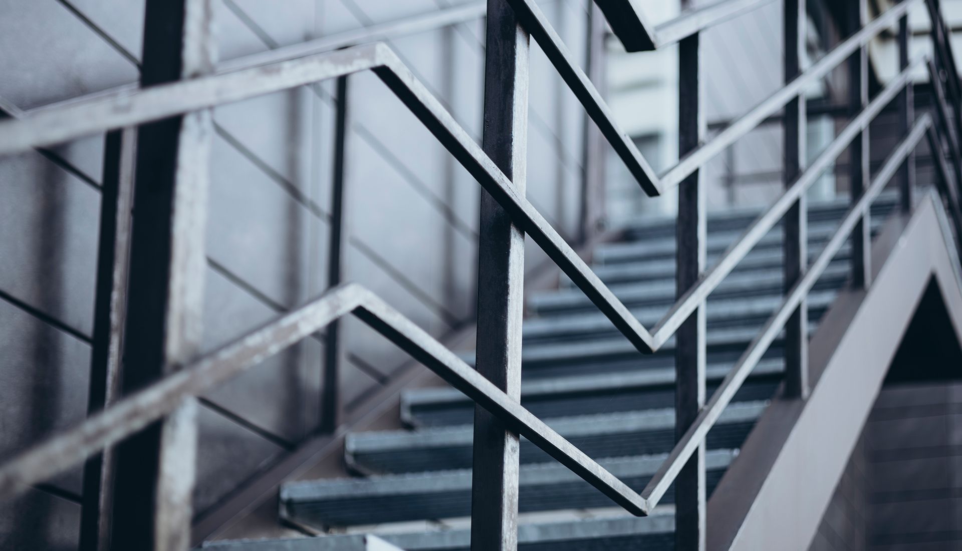 Metal staircase with zigzag railing, leading upwards. Grey and blue tones.