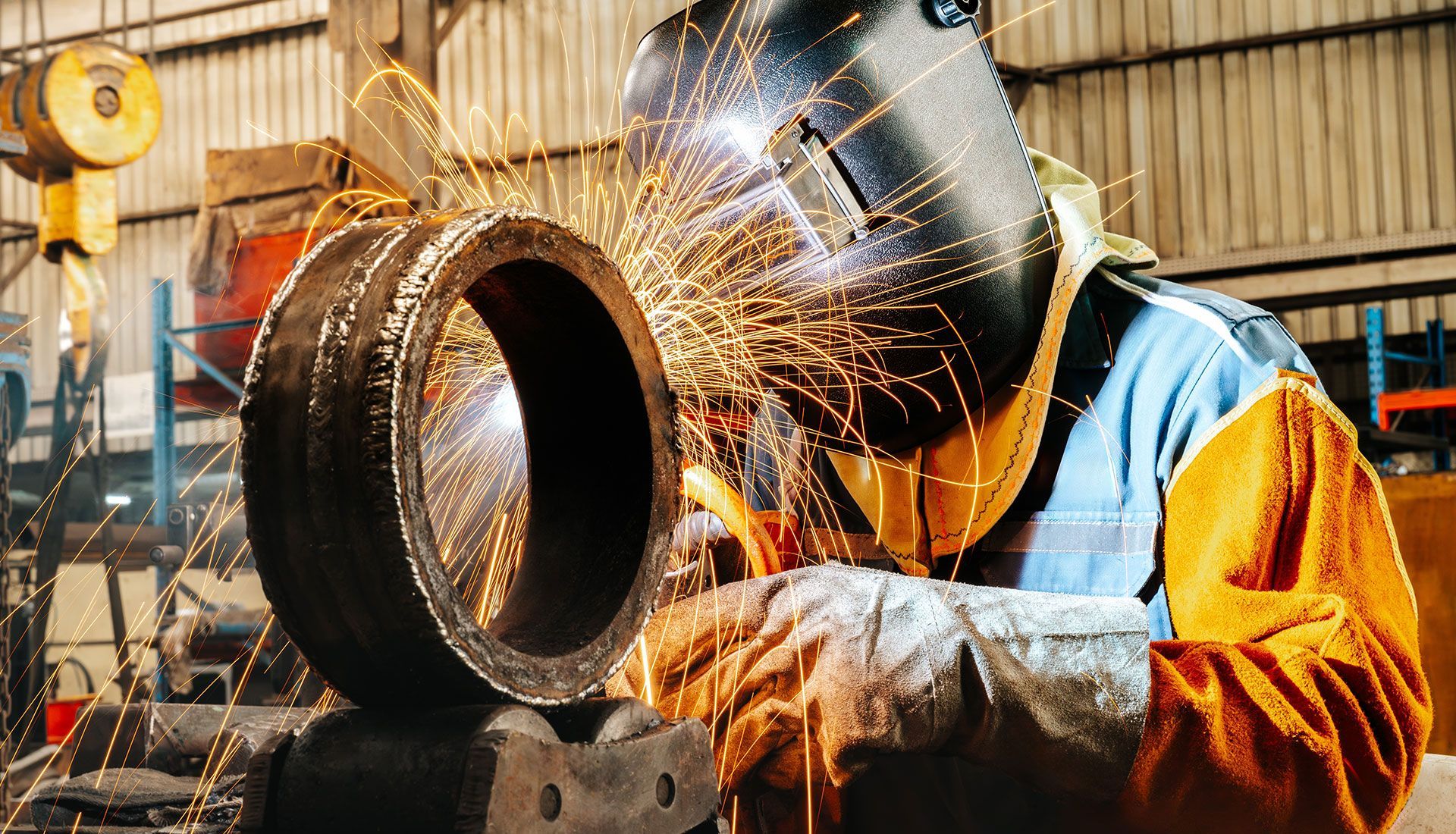 Welder in protective gear welding a large, circular metal object in a workshop. Sparks fly.