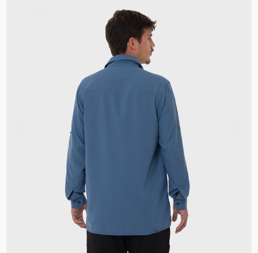 Hombre con camisa azul abotonada con las mangas remangadas y bolsillo en el pecho, de pie contra un fondo blanco.