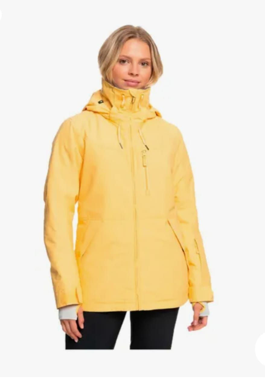 Mujer con chaqueta de esquí amarilla, de pie frente a un fondo blanco.