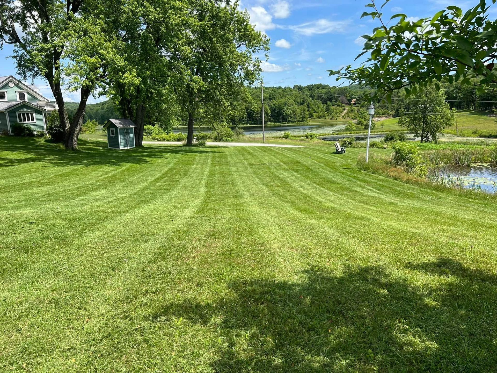 Lawn with fresh mowing stripes beside a pond, trees, and a house in a sunny rural yard