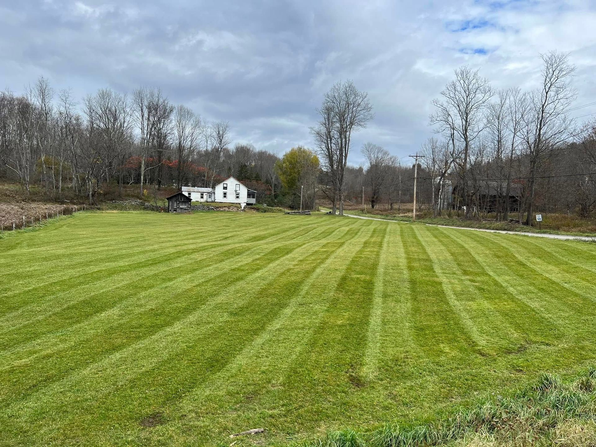Wide grassy field with striped mowing lines, a small white house, and leafless trees under a cloudy sky.