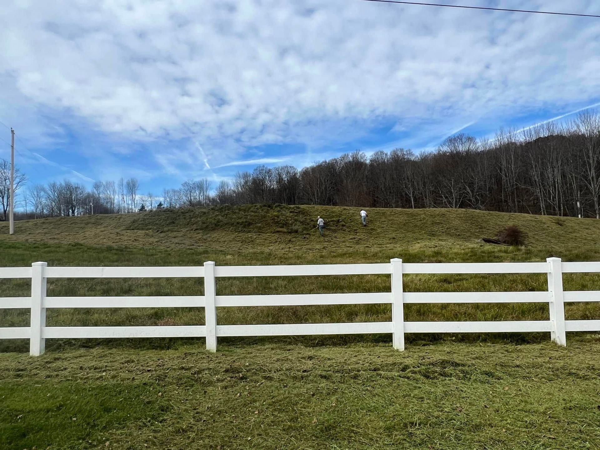 White fence in a grassy field with a wooded hill and cloudy blue sky behind it.