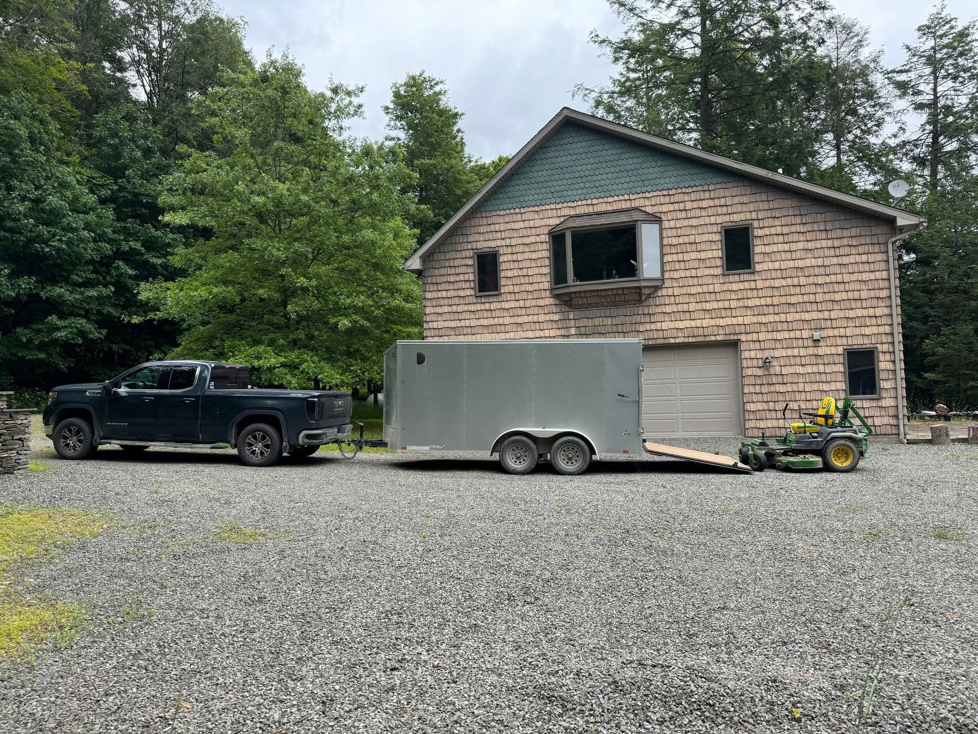 Black pickup truck and gray utility trailer parked in front of a stone house with garage.