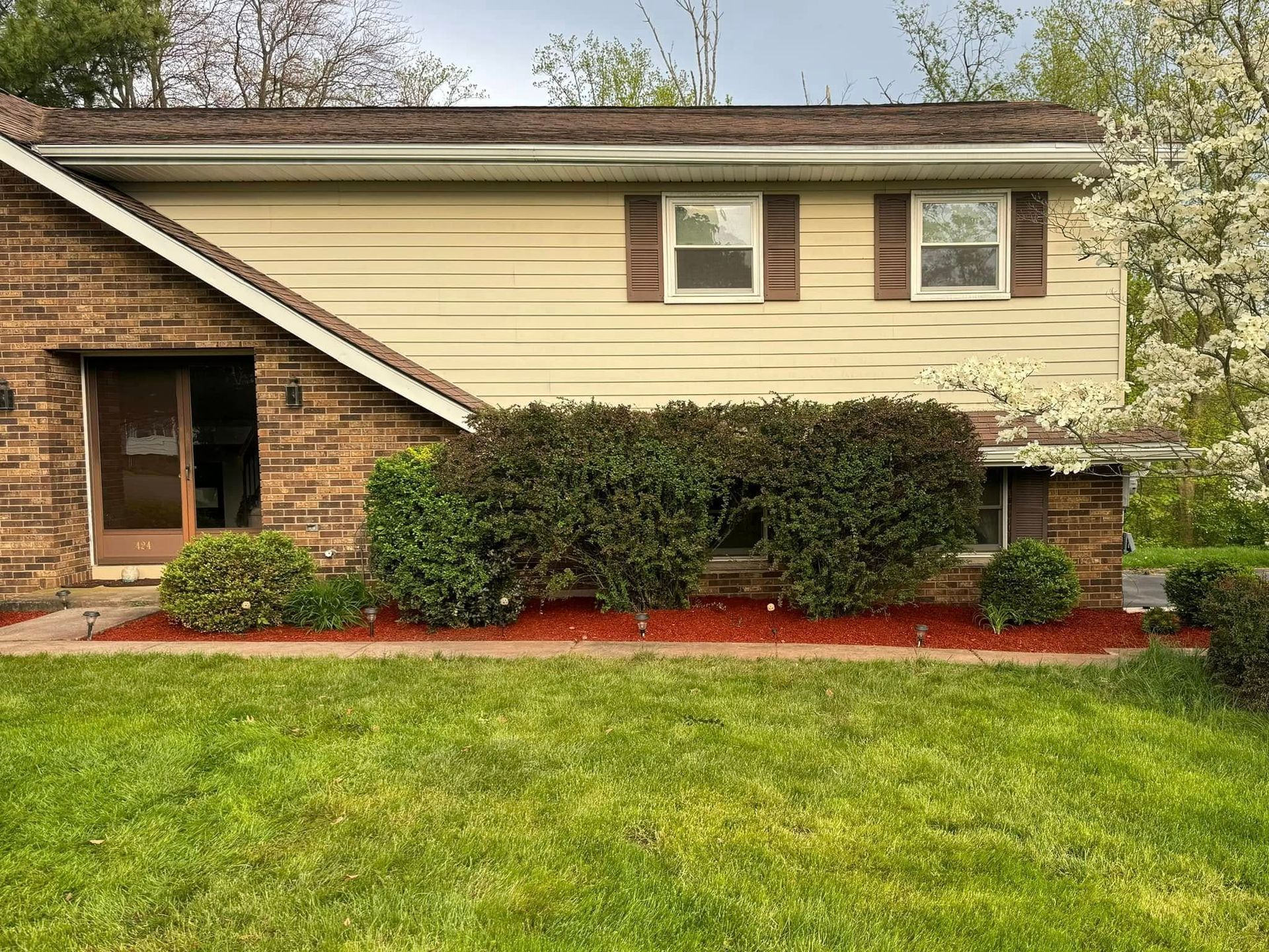 Suburban house exterior with tan siding, brick entry, shrubs, and a green front lawn.