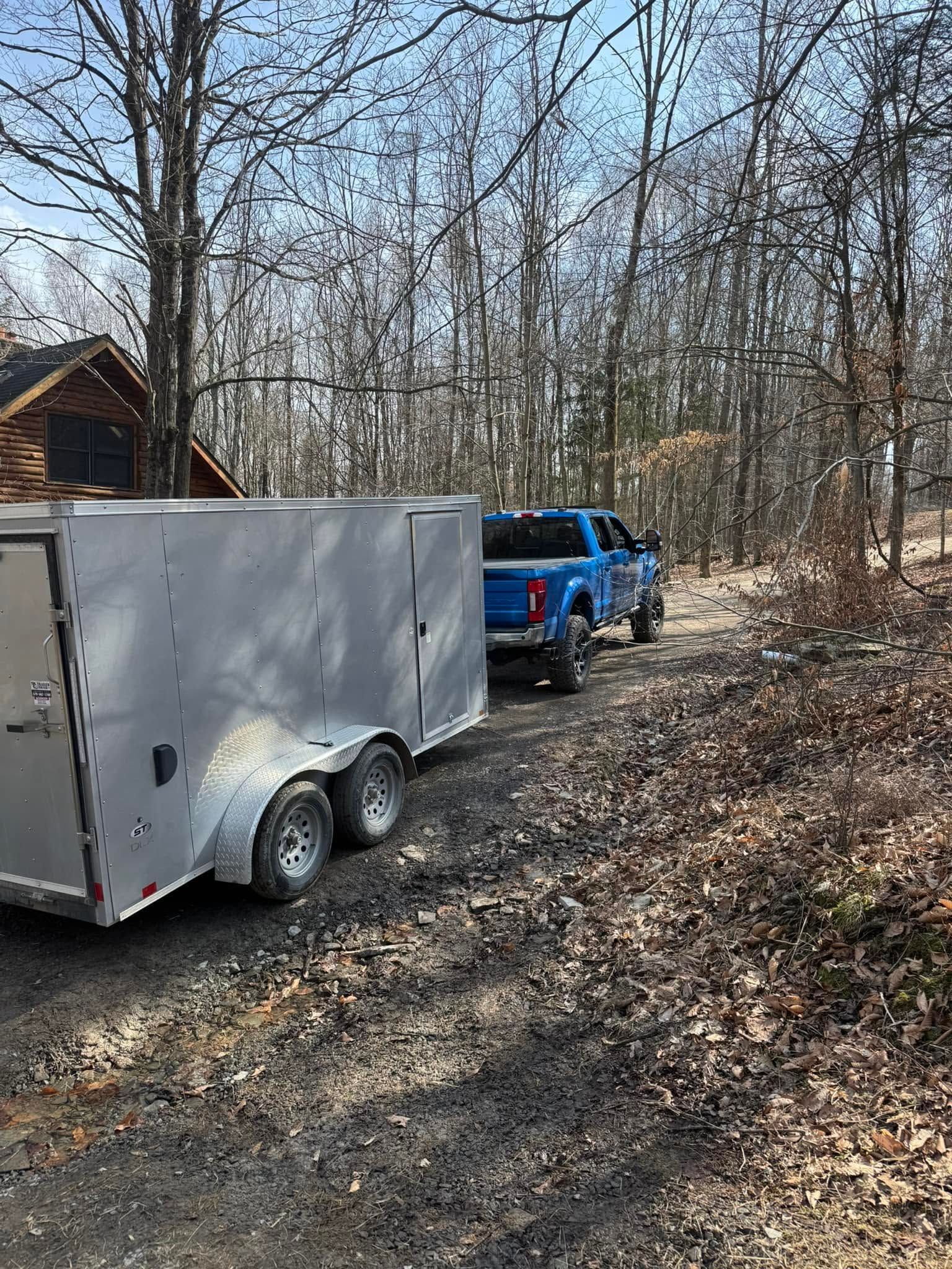 Blue pickup towing a white trailer on a wooded dirt road beside a small cabin