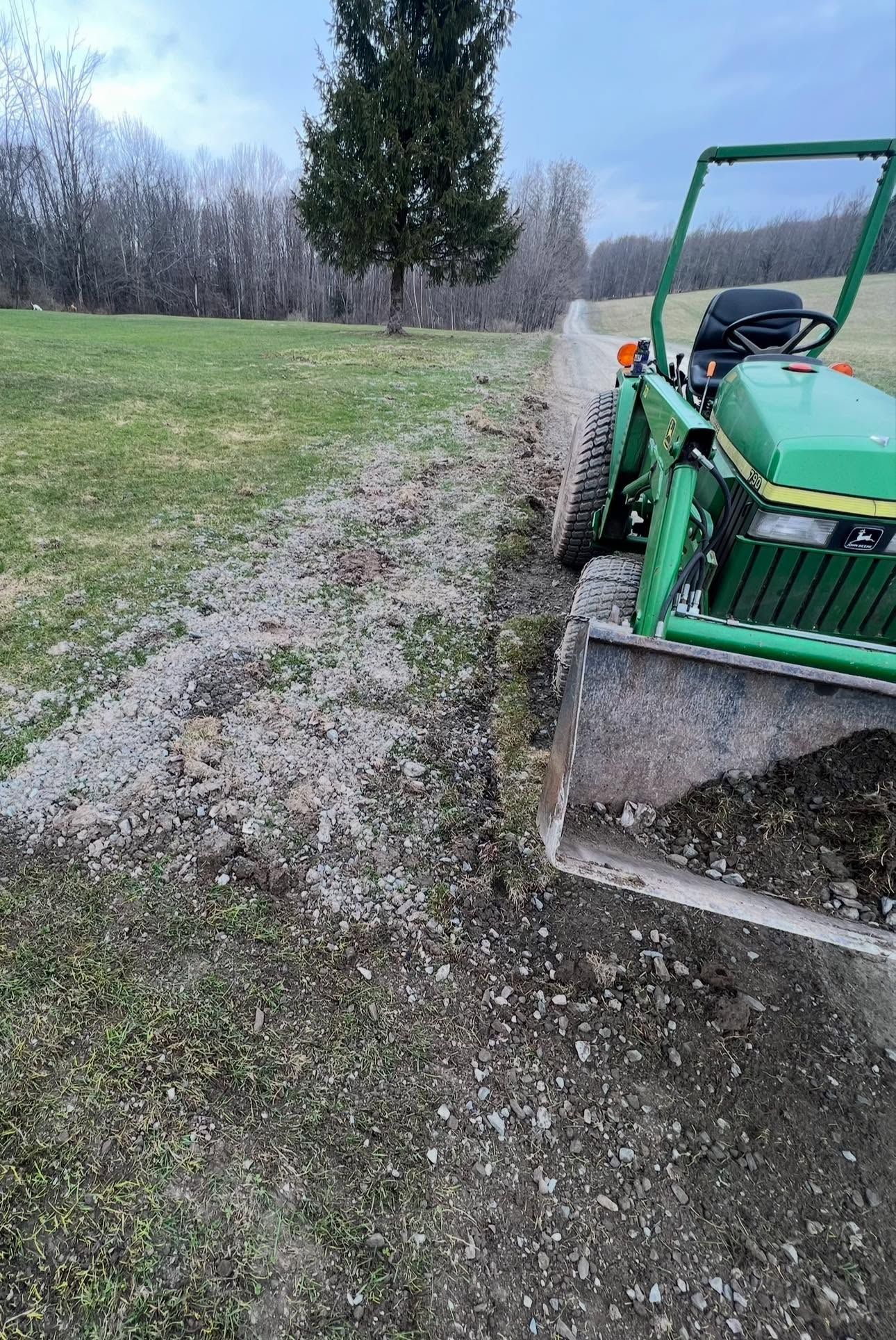 Green tractor with front blade on a gravel path beside a grassy field and trees under a cloudy sky