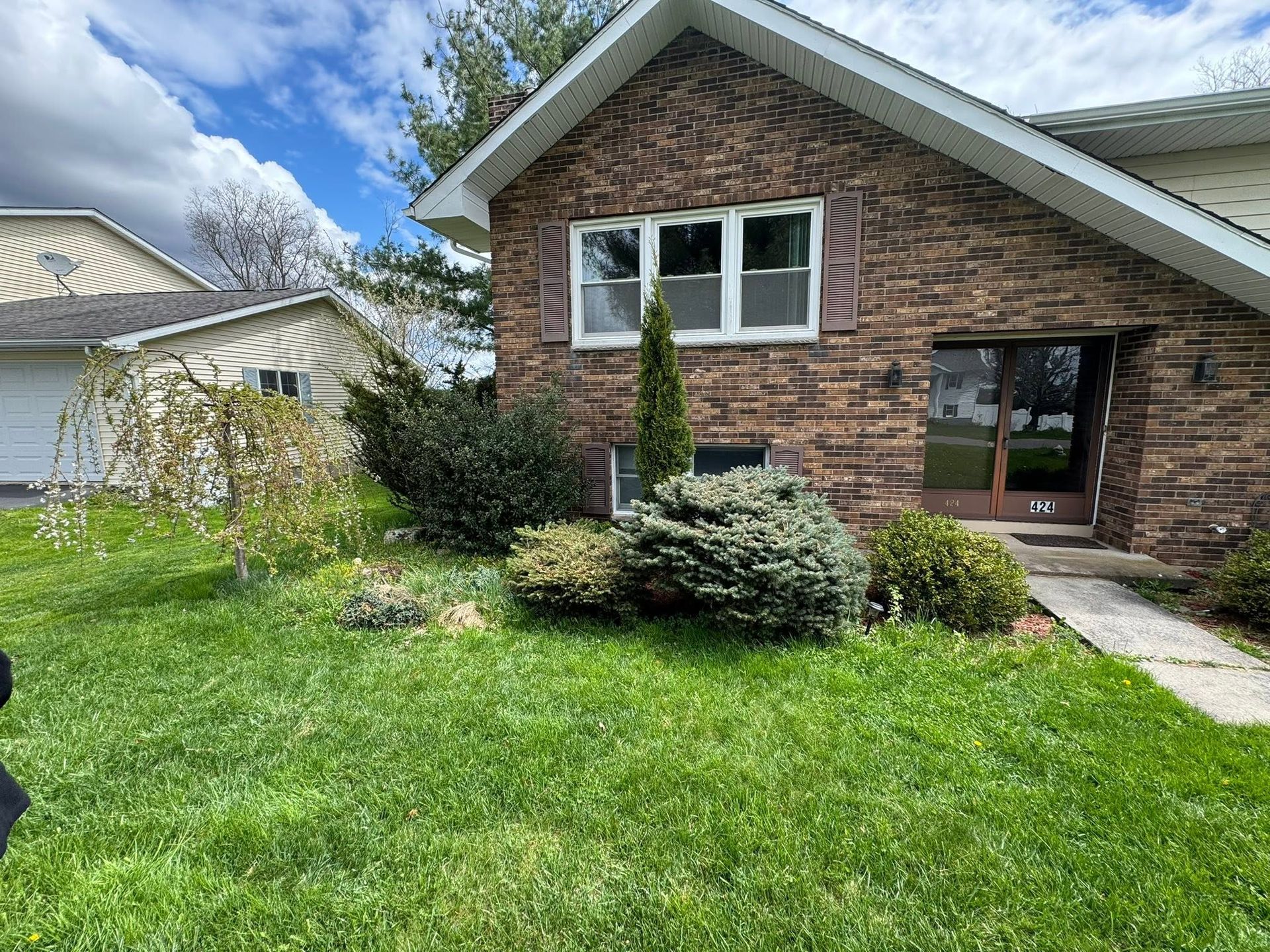 Suburban house exterior with brick and siding, green lawn, shrubs, and a front walkway under a blue sky