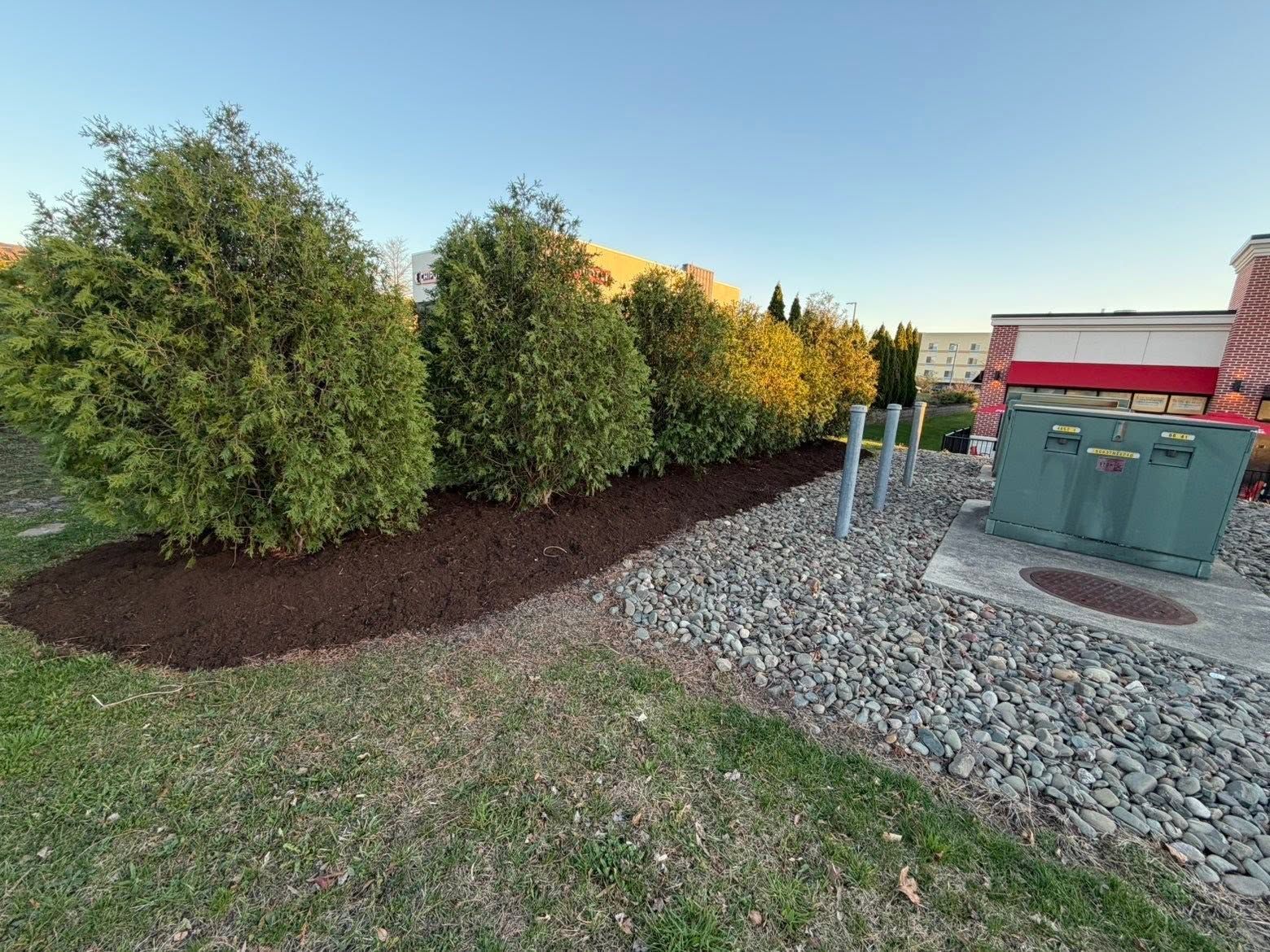 Shrubs along a gravel and mulch landscape beside utility boxes and a red building.