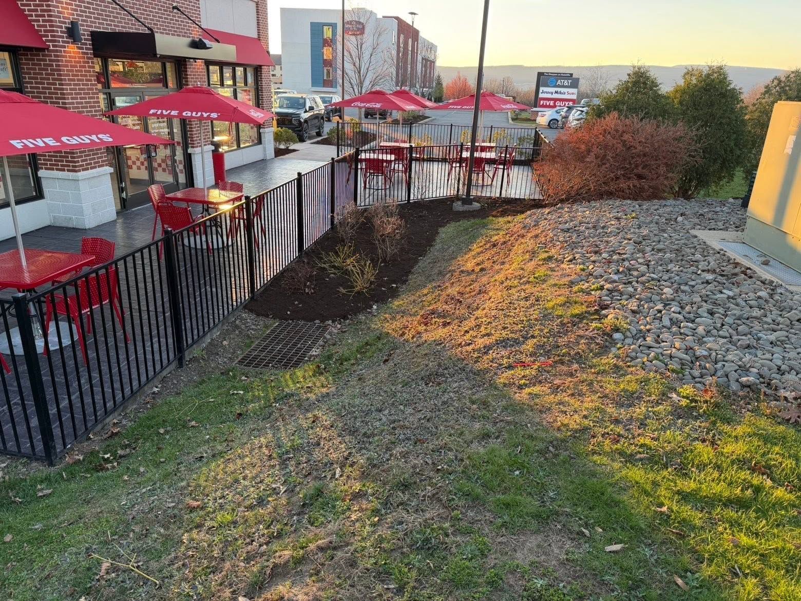 Outdoor patio beside a building, with red umbrellas, black railings, and a rocky landscaped slope at sunset