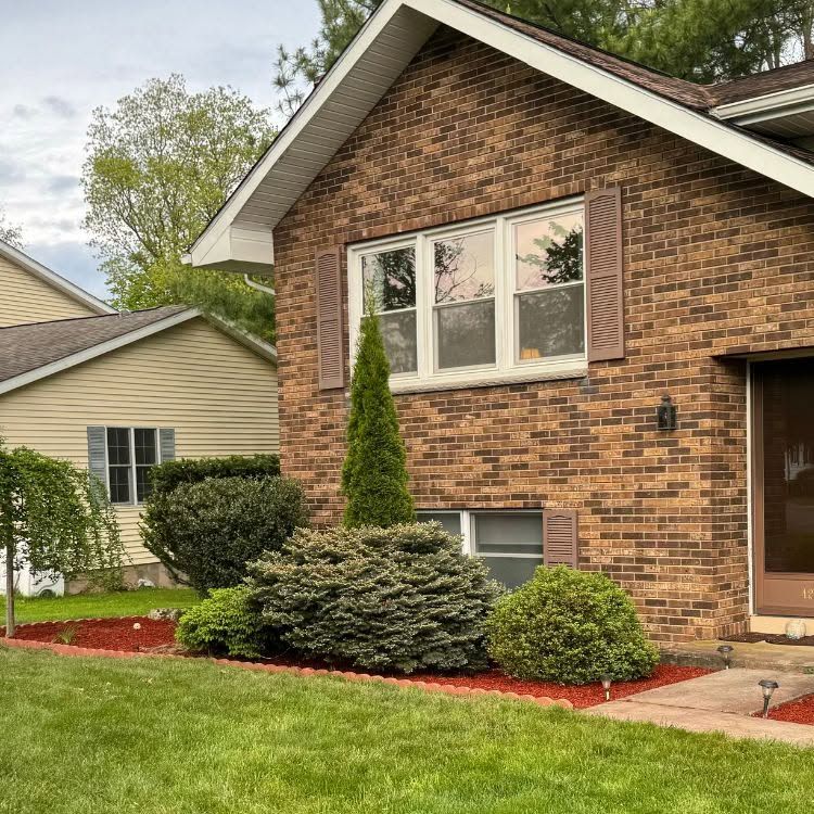 Brick suburban house with white-trim windows, brown shutters, and a manicured lawn and shrubs.