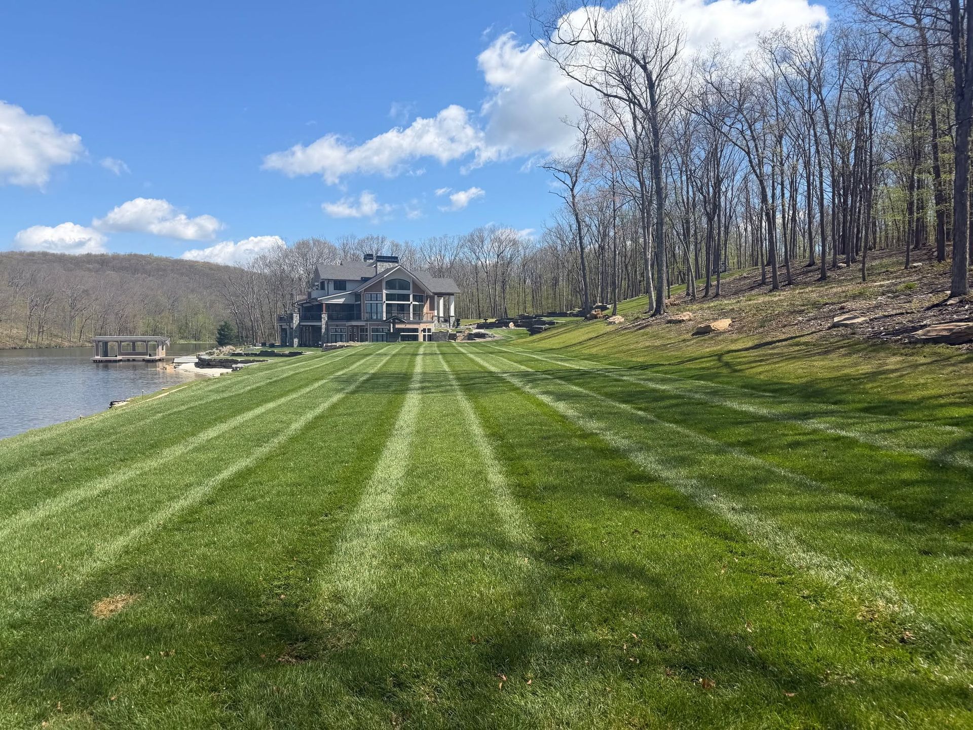 Lakeside lawn with striped grass, a house in the distance, and trees under a blue sky with clouds