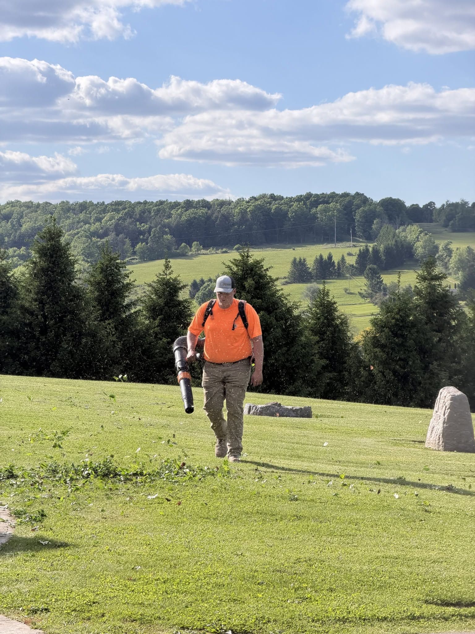 Hikers in orange and camo walk through a grassy field with trees and hills under a partly cloudy sky