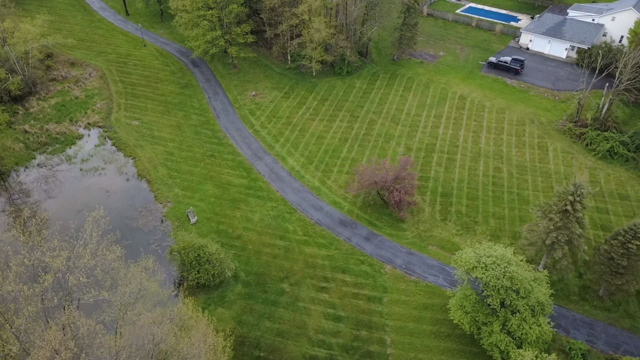 Aerial view of a grassy field beside a pond, with a winding path and a house at the top right.