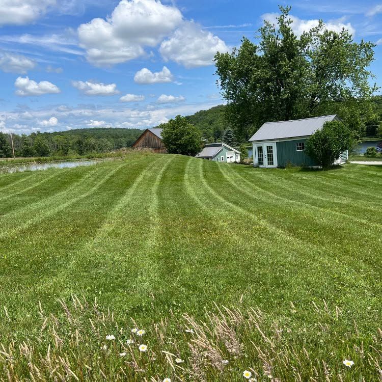 Wide mowed green lawn with a small white house, shed, trees, and cloudy blue sky