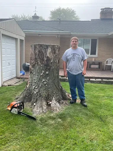 A man is standing next to a tree stump in front of a house.