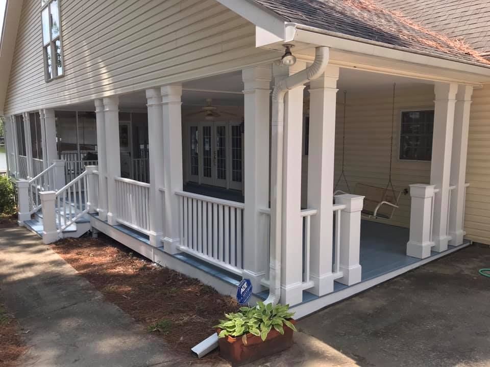 White-columned porch with railing, next to a tan house. Blue porch floor, green plants, paved driveway.