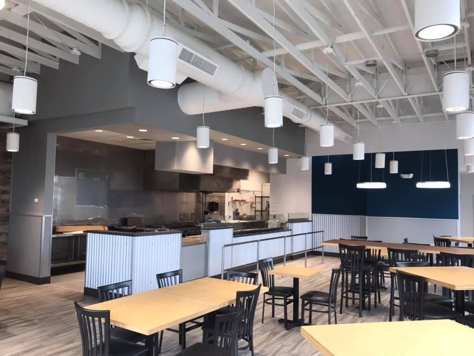 Interior of a cafeteria with tables, chairs, and a kitchen area. Overhead lights and air ducts are visible.