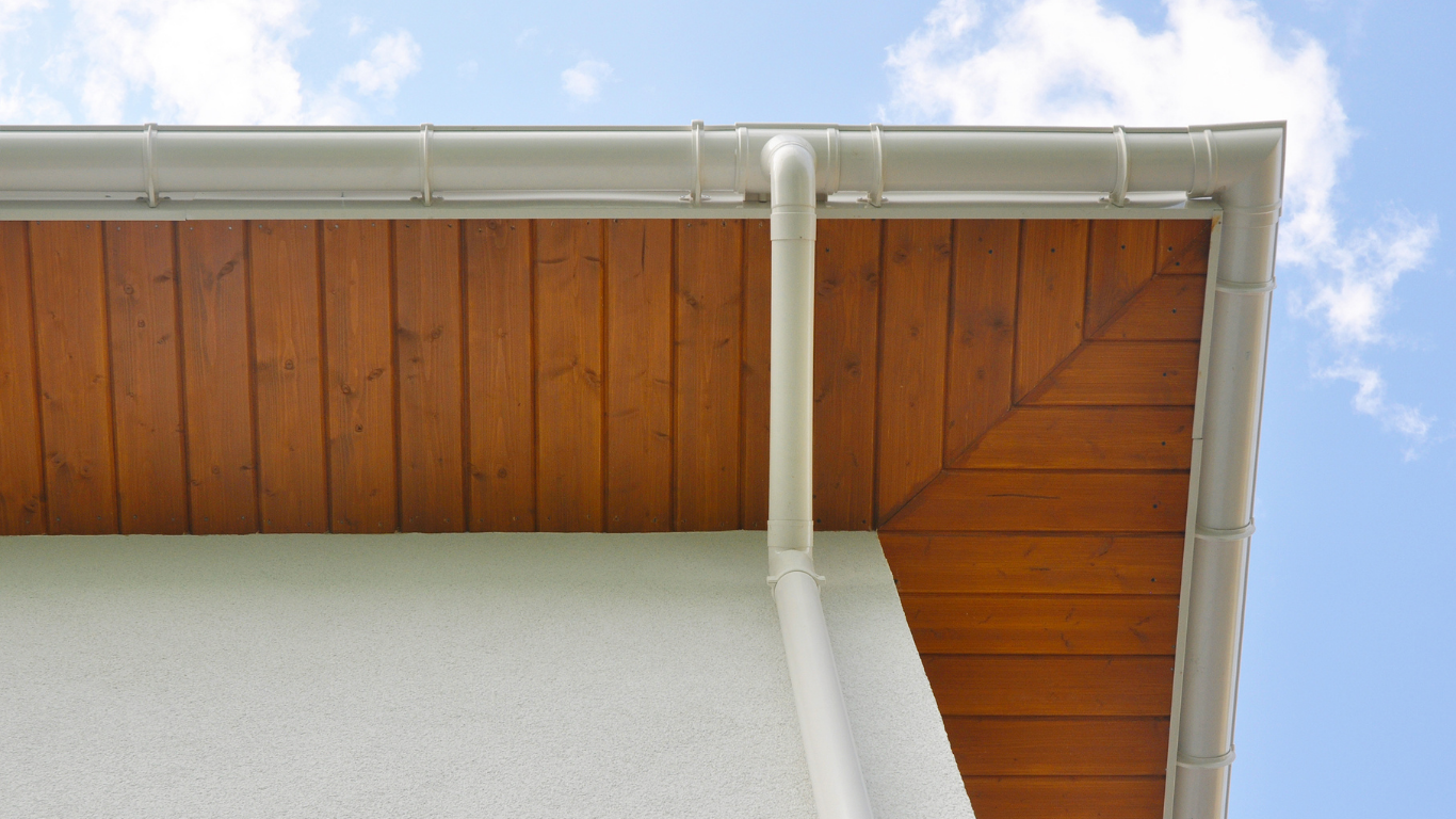 White gutters and downspout on a home with a wooden soffit and a blue sky background.