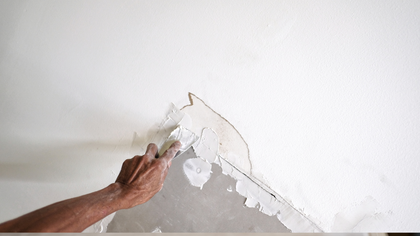 Person scraping peeling white paint from a wall with a putty knife.
