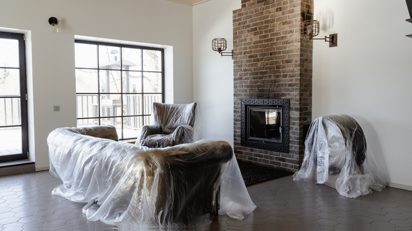 Living room with furniture covered in plastic, fireplace, and large windows.