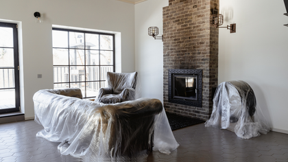 Living room with furniture covered in plastic, fireplace, and large windows.