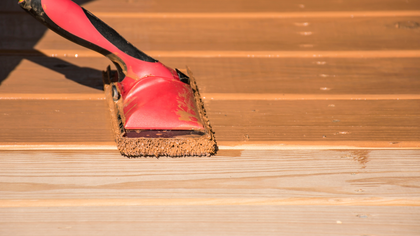 Painting a wooden deck with a stain. A red applicator applies the stain to the planks, showing the before and after.
