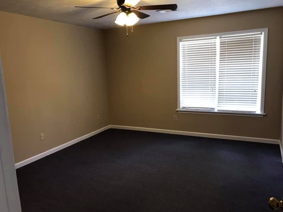 Empty bedroom with beige walls, dark carpet, and a window with closed blinds.