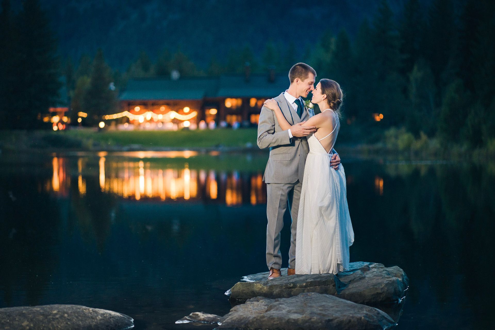 Couple embraces on a rock in a lake at night; a building with lights reflects in the water.