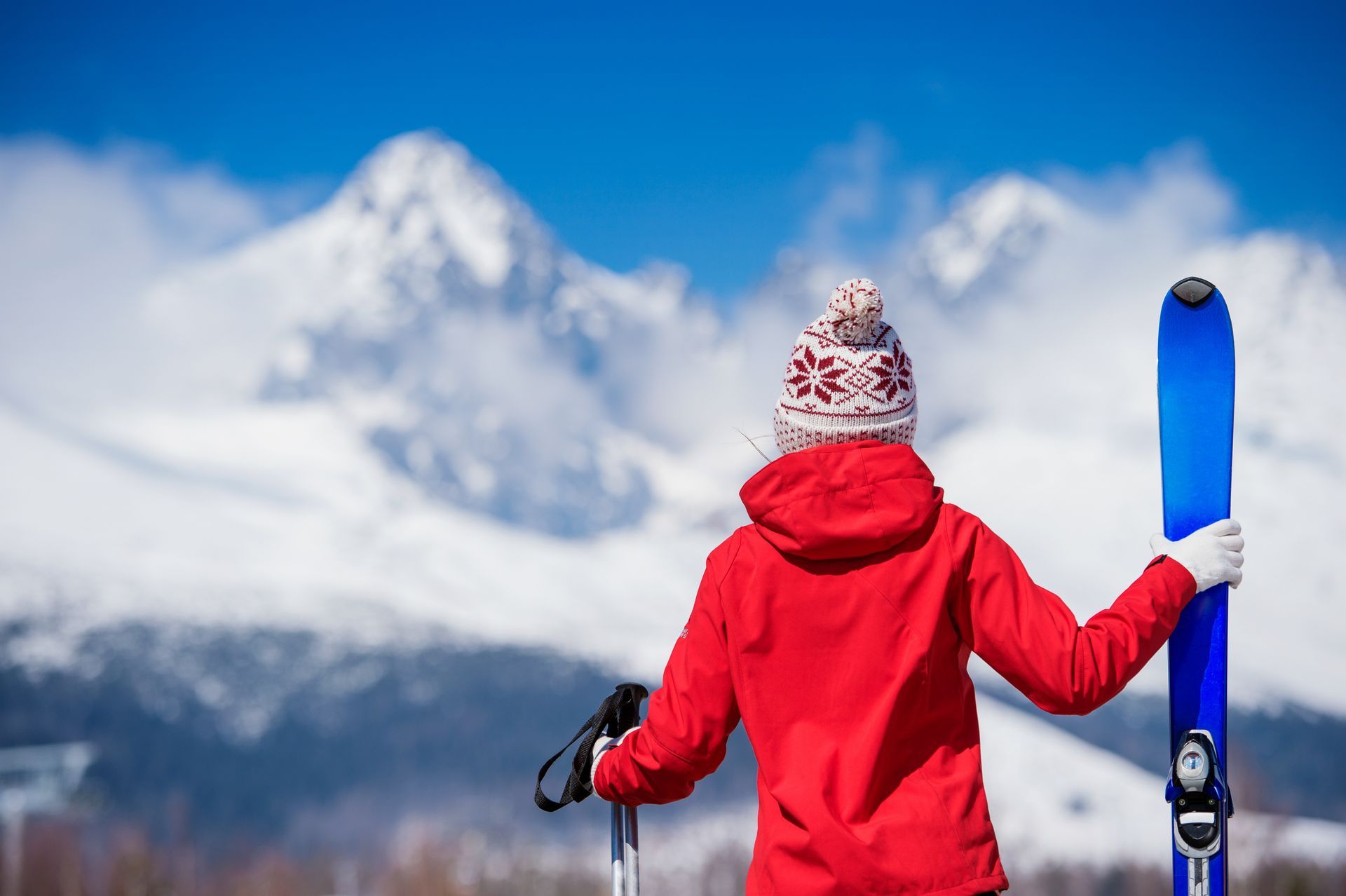 Person in red jacket and hat holding skis, facing snowy mountains, clear blue sky.